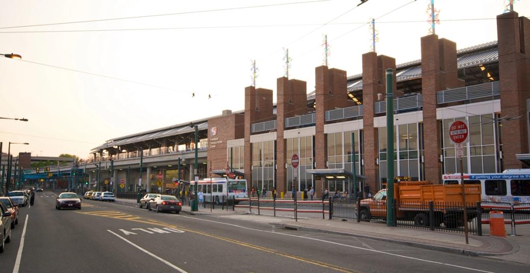 Brick building with large glass windows connected to an elevated train line. On the street level, several buses are idling as passengers board while cars drive down the avenue.