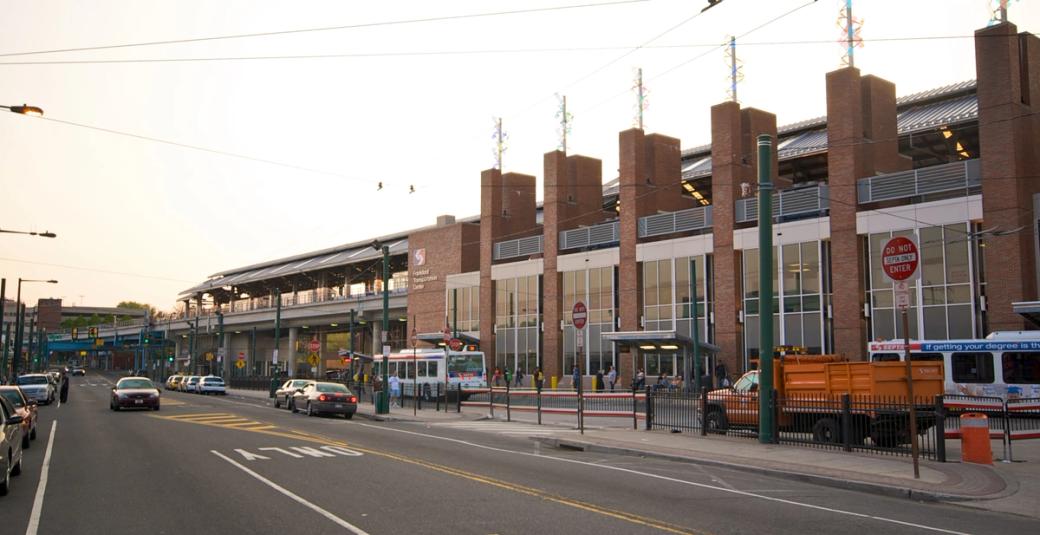 Brick building with large glass windows connected to an elevated train line. On the street level, several buses are idling as passengers board while cars drive down the avenue.