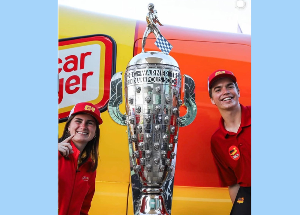 Two people in red uniforms standing next to a large silver trophy.