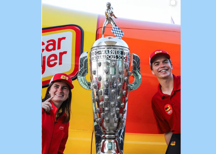 Two people in red uniforms standing next to a large silver trophy.