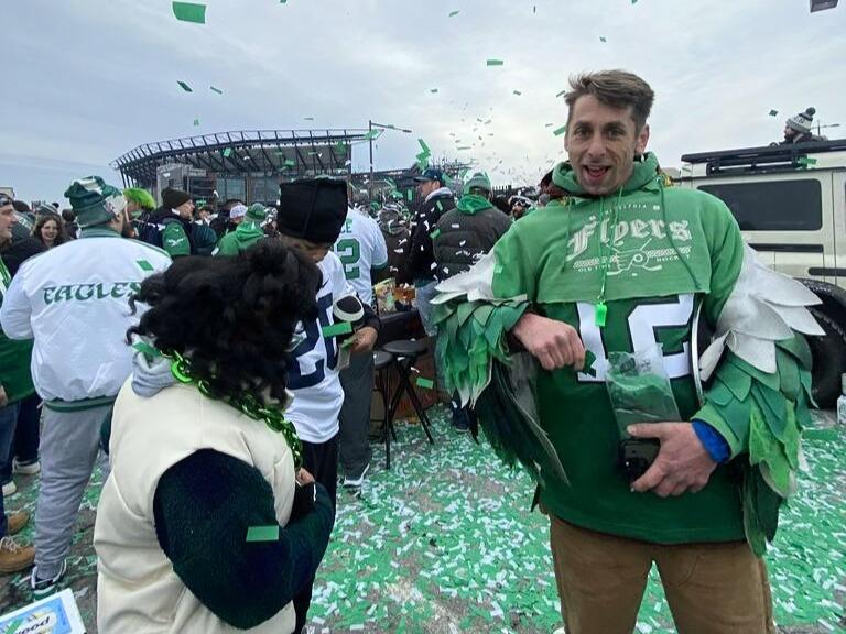 Green confetti flies through the air and litters the ground on an overcast gameday. Fans are dressed in Eagles gear. To the right, a smiling fan wears a green Eagles Jersey with a Flyers sweatshirt cut off with added wings.