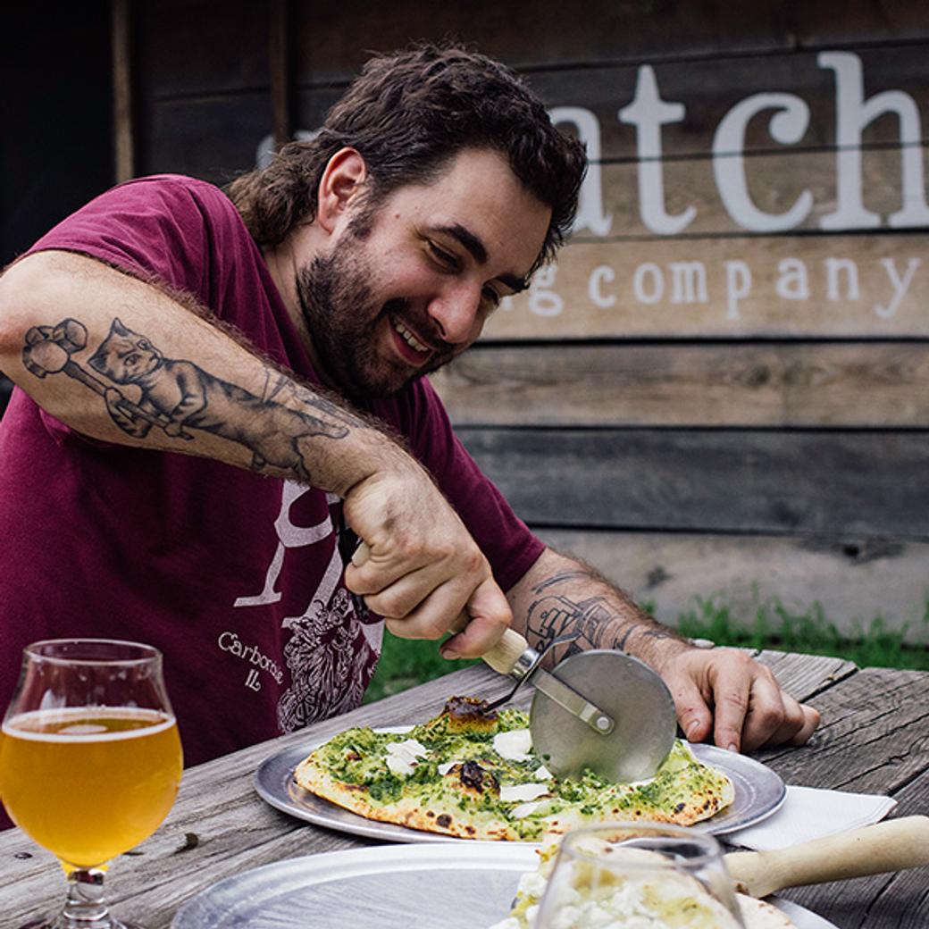 A man with tattoos, wearing a maroon shirt, smiles while cutting a pizza with a pizza cutter. A glass of beer is on the wooden table outdoors.