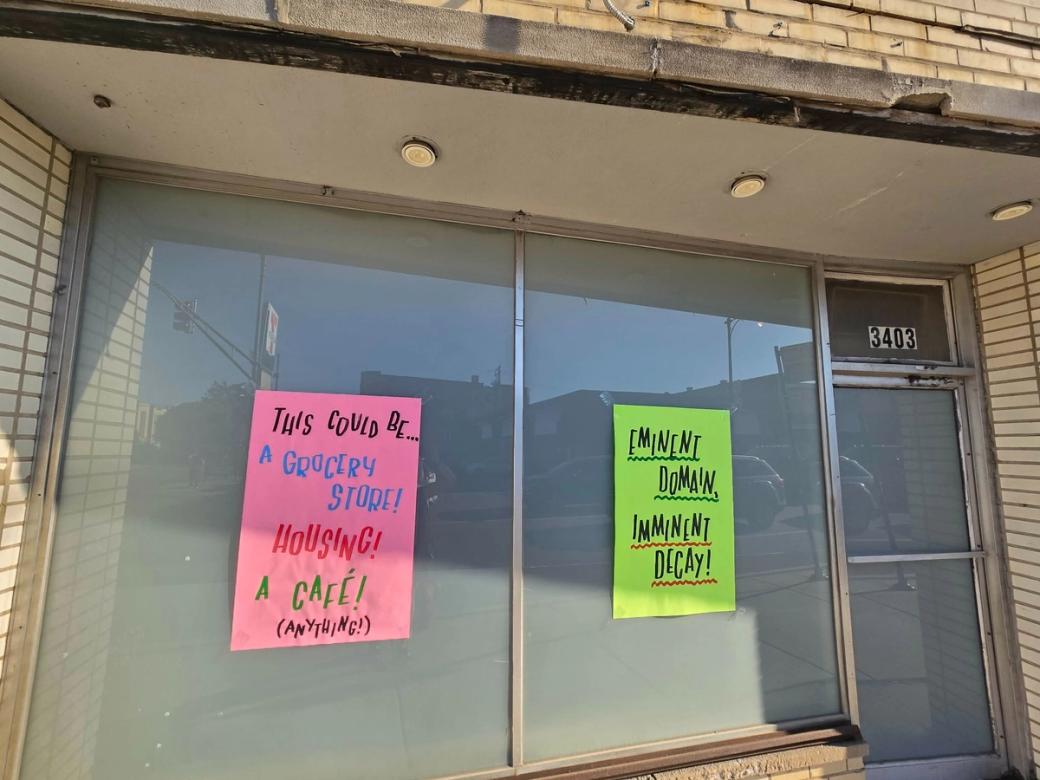 Two signs on a window of an abandoned building. They read "this could be a grocery store, housing, cafe" the other sign reads "eminent domain, imminent decay"