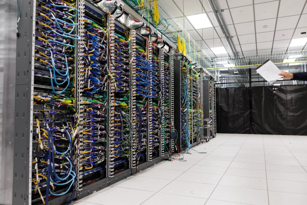 A wall of servers in a room with a person’s arm holding a legal pad gesturing towards the servers. 