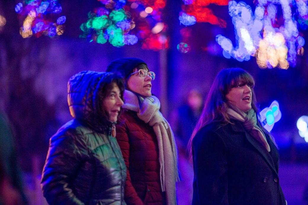 Three people wearing jackets look up at blurred holiday lights