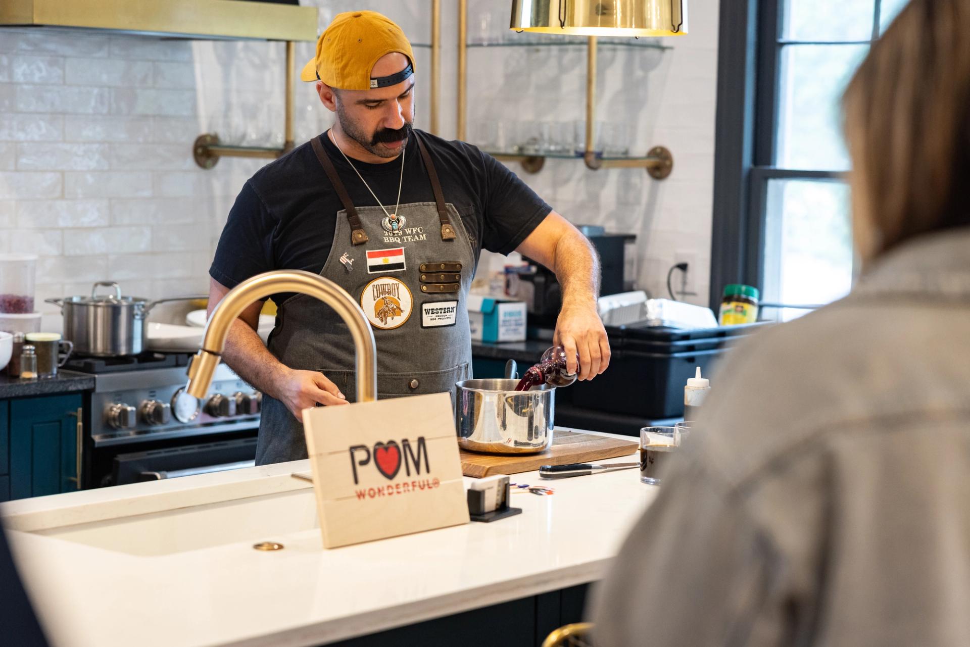 A man wearing a black T-shirt and a gray apron stands in a kitchen and pours pomegranate juice into a silver pot.