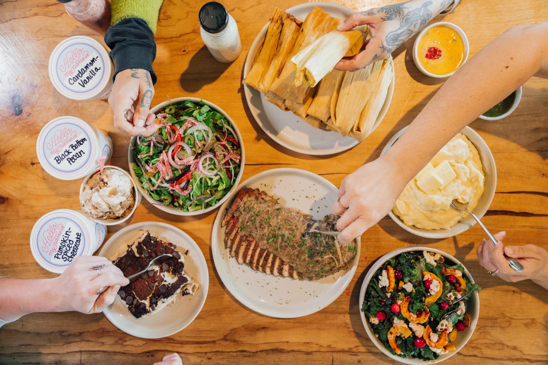 A wooden table covered with plates. The plates are holding meat, salad, tamales, mashed potatoes, and chocolate cake. Hands reach over the table to dish out food. Pints of ice cream are nearby.