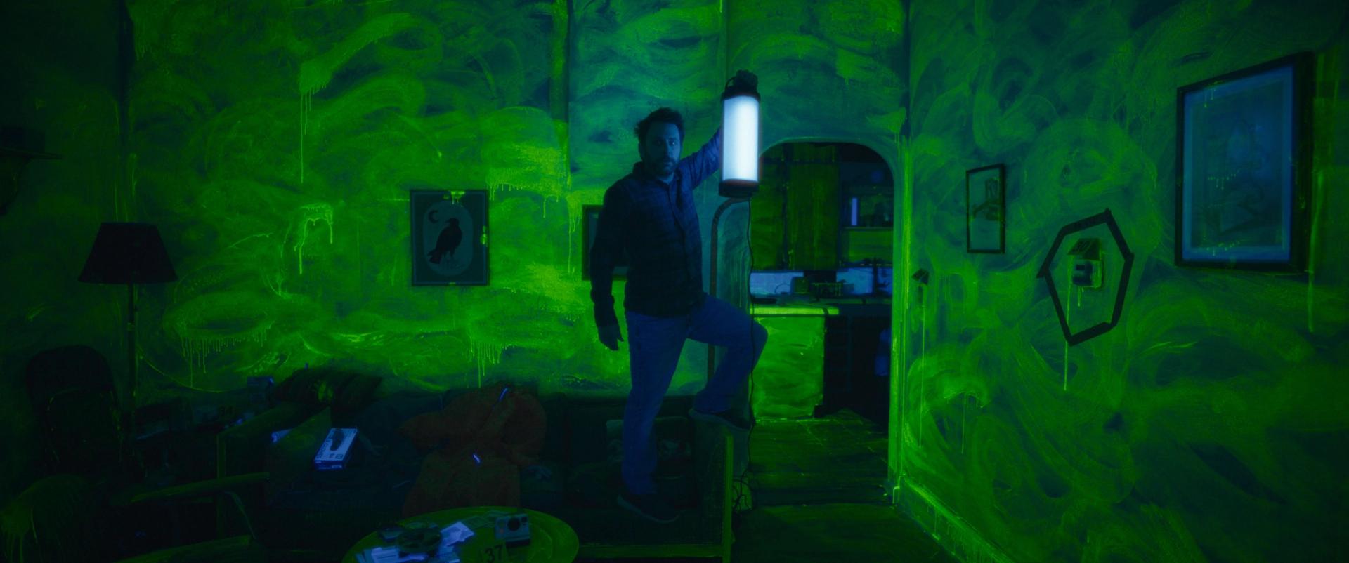 Charlie Day holding a black light in a neon green room. 