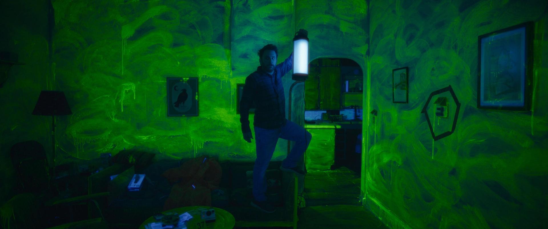 Charlie Day holding a black light in a neon green room.