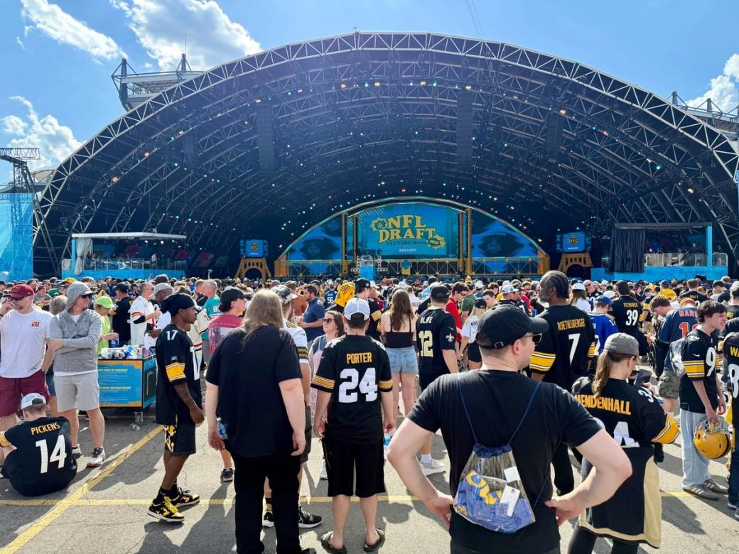 A large crowd of football fans, many wearing Pittsburgh Steelers jerseys including numbers 14 (Pickens), 24 (Porter), 7 (Roethlisberger), and 34 (Mendenhall), gathered in front of the main stage at the 2025 NFL Draft in Pittsburgh. The stage displays a large 'NFL Draft Pittsburgh' logo on LED screens under an open-air arched structure on a sunny day.