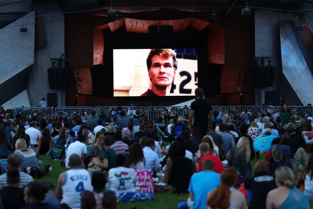 A crowd of people sitting in a field watching a move projecting on a screen