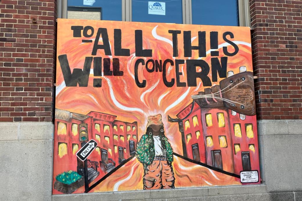 A mural on a square piece of wood designed like an album cover. Bold, black text says “To All This Will Concern” at the top. A faceless Black woman wearing an orange hat, white shirt, green puffy jacket, and baggy orange pants stands in the center of a street with rowhomes on either side, with the backdrop painted in hues of orange, white, yellow, red, and brown.