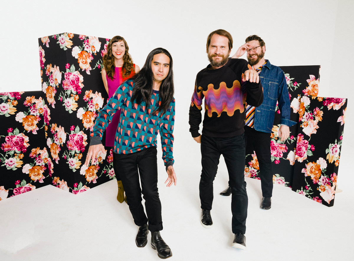 Three men and a woman pose in front of black boxes covered in a flower print.