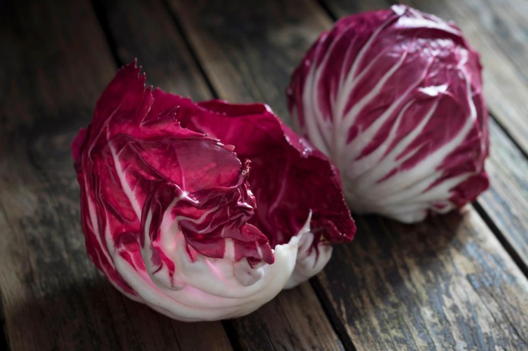 A loose leaf and full head of radicchio sit on a wooden table. The vegetable’s leafy ends are bright magenta with white veins.