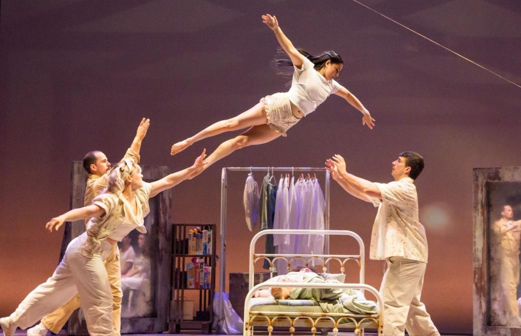 Four people on stage, there are two on the left that look to be throwing someone through the air to the person on the right. The stage looks to have a bed and a closet behind it