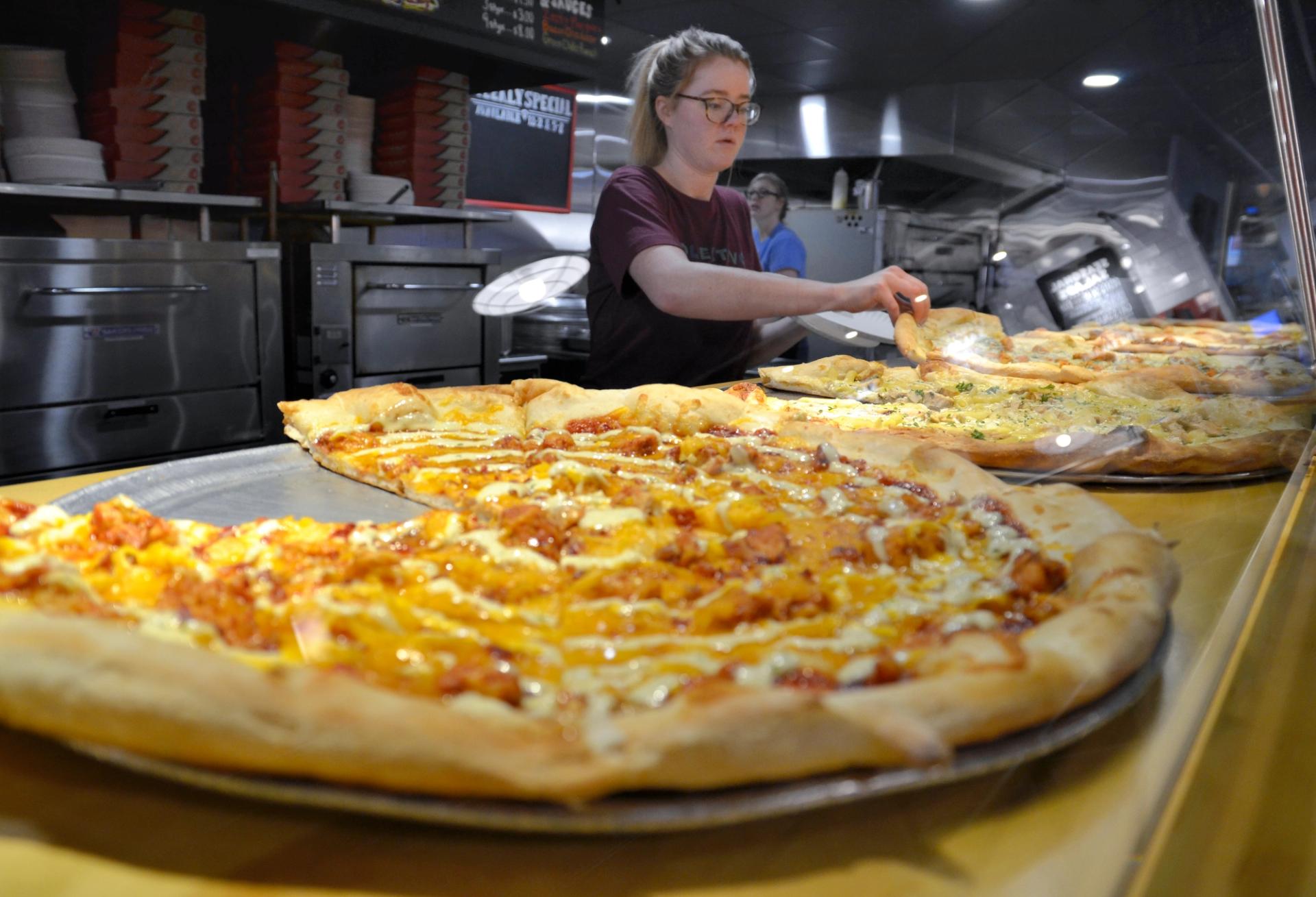 A woman stands behind a row of pizzas.