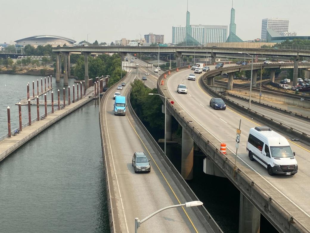Cars on multiple highway lanes next to a river with buildings in the distance.