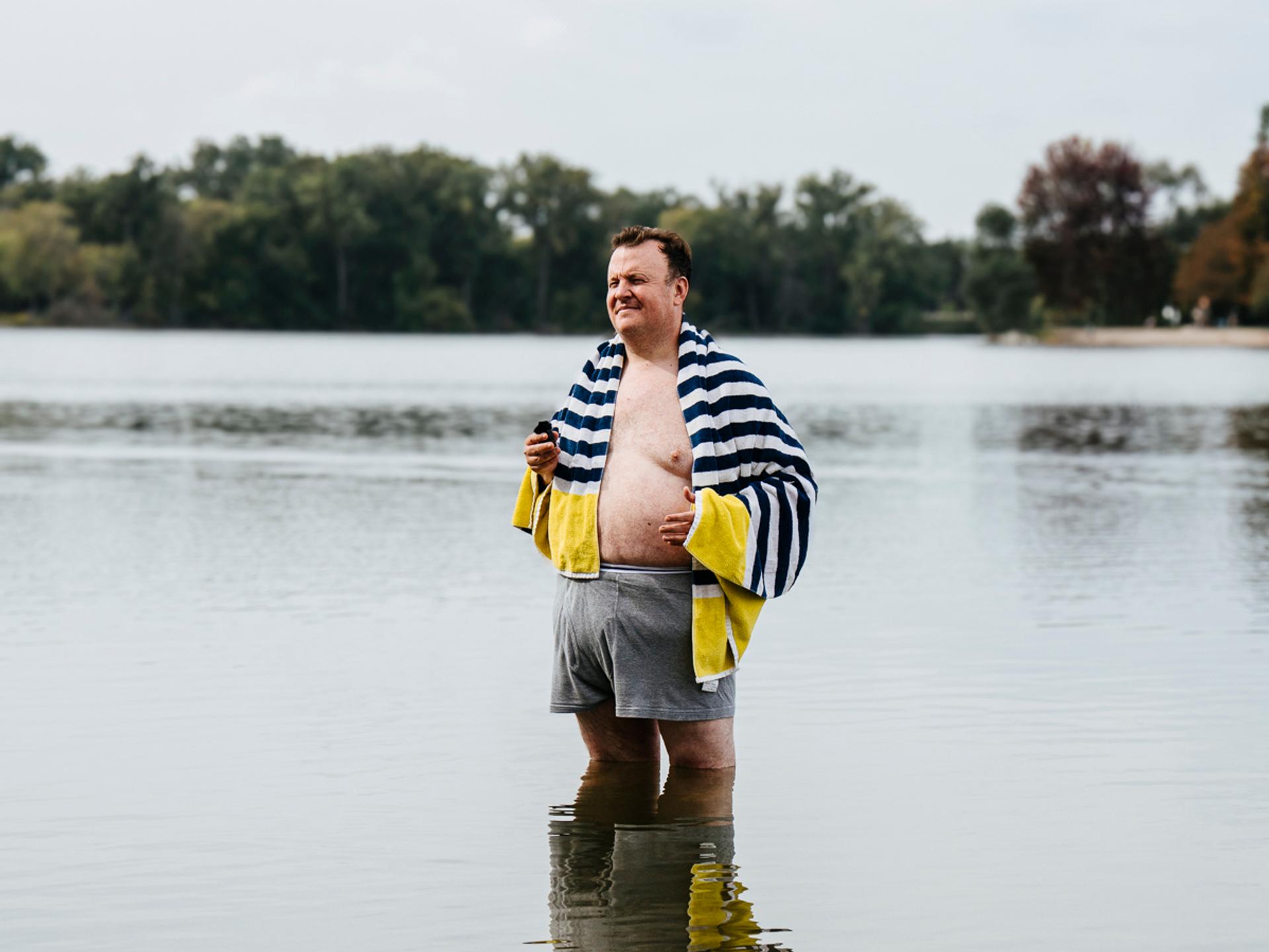 A man with a towel on his shoulders standing in a lake. There are trees in the background as well