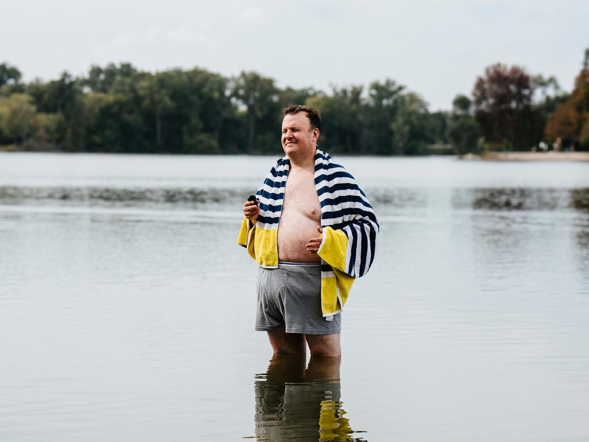 A man with a towel on his shoulders standing in a lake. There are trees in the background as well