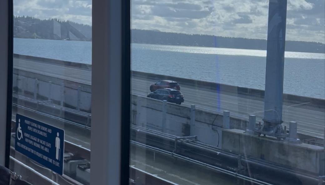 The view from inside Sound Transit’s 2 Line crossing Lake Washington on the floating I-90 bridge. Out the window cars can be seen driving across the bridge’s span with the lake and a blue sky and puffy white clouds in the background.