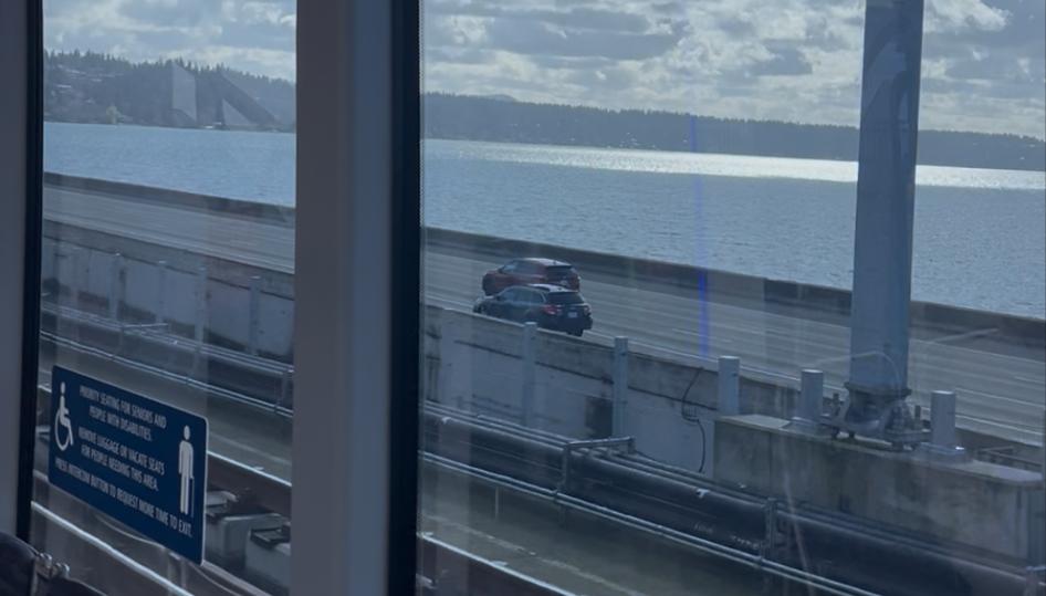 The view from inside Sound Transit’s 2 Line crossing Lake Washington on the floating I-90 bridge. Out the window cars can be seen driving across the bridge’s span with the lake and a blue sky and puffy white clouds in the background.