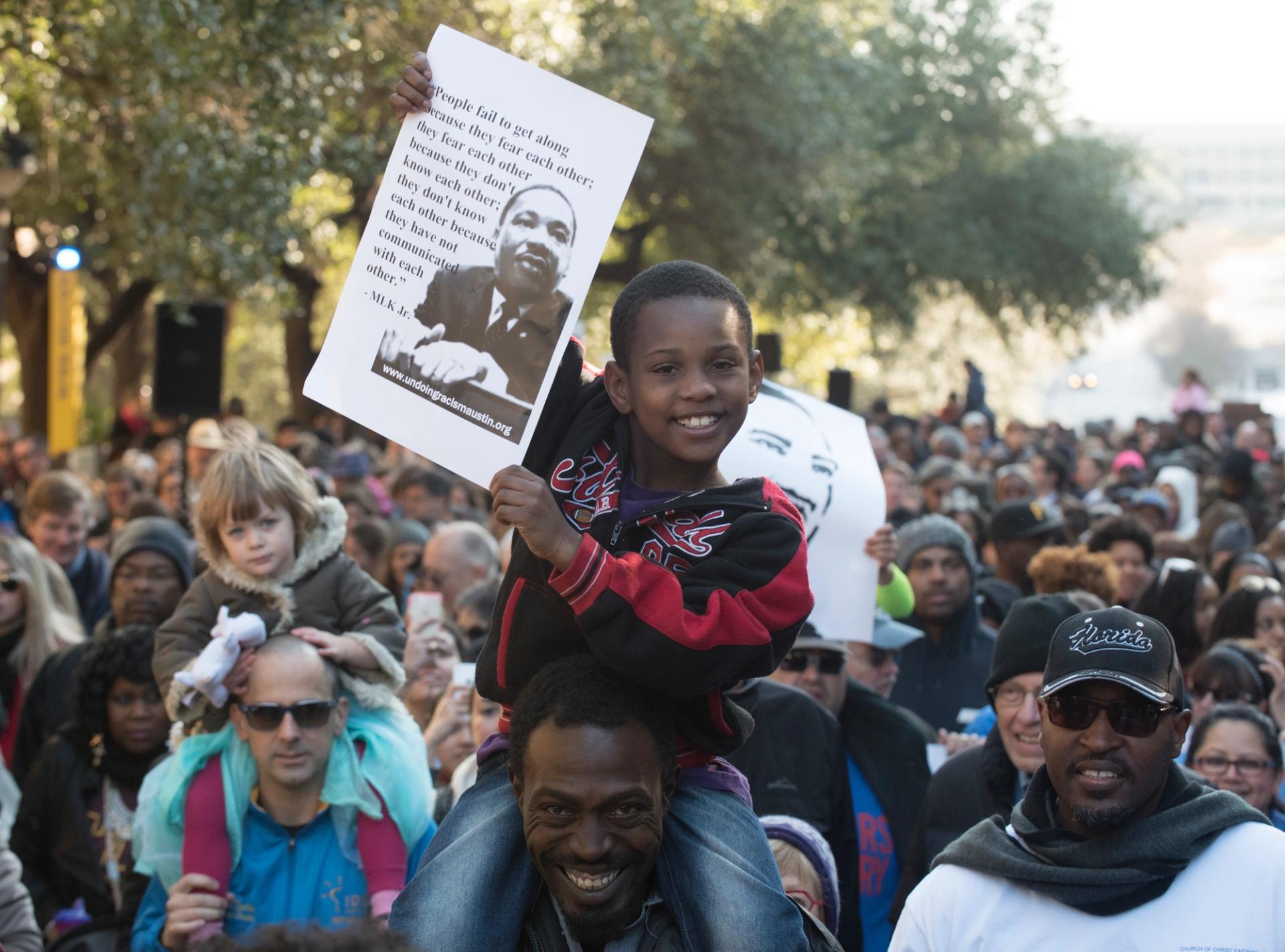 A boy holds up a Martin Luther King, Jr. poster while sitting on the shoulders of a man. The pair are part of an MLK Day crowd.