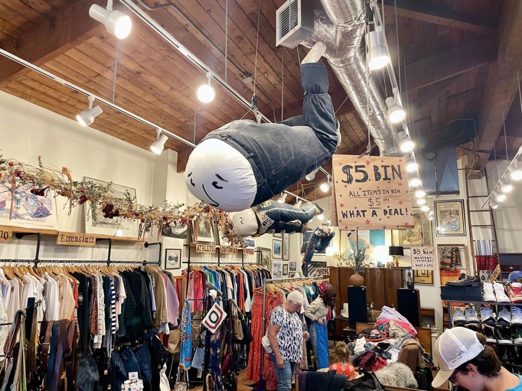A packed second-hand clothing store with people shopping, multi-colored clothing on racks, and art on the walls and hanging from the ceiling.