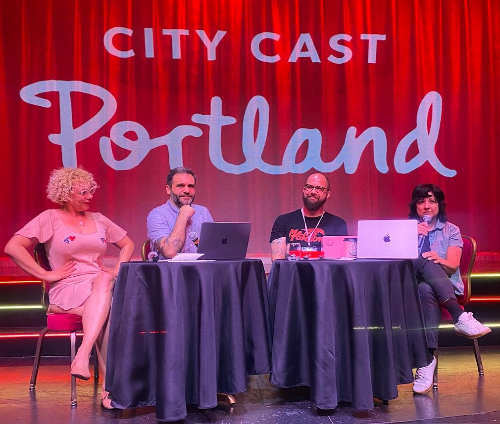Four people sit at two tables on stage with red curtain behind them. Laptops sit on each table.