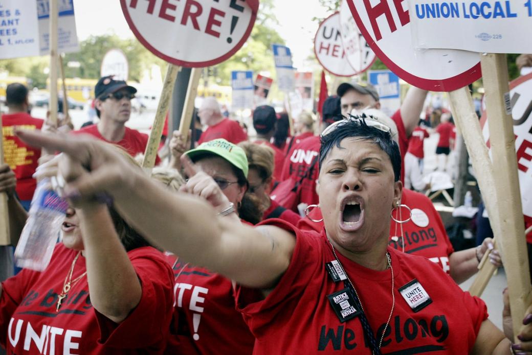 A group of people in red shirts that say "we belong unite" protesting. A woman in the foreground seems to be mid chant and pointing