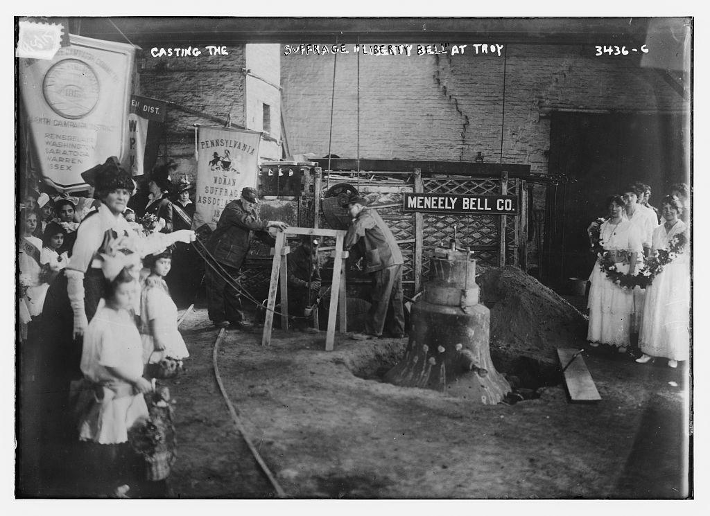 A black and white photo of women standing in a room with a bell. There is a sign that reads "MENEELY BELL CO."