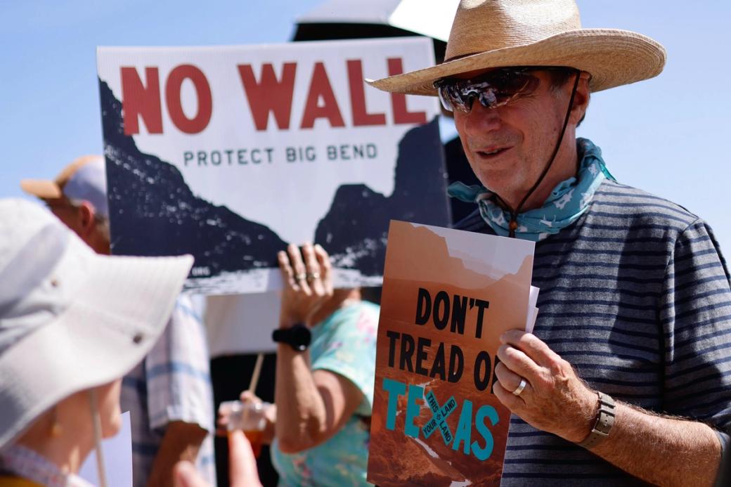 A man wearing a straw hat and holding a sign that says "Don't tread on Texas." A sign behind him says "No wall protect Big Bend."