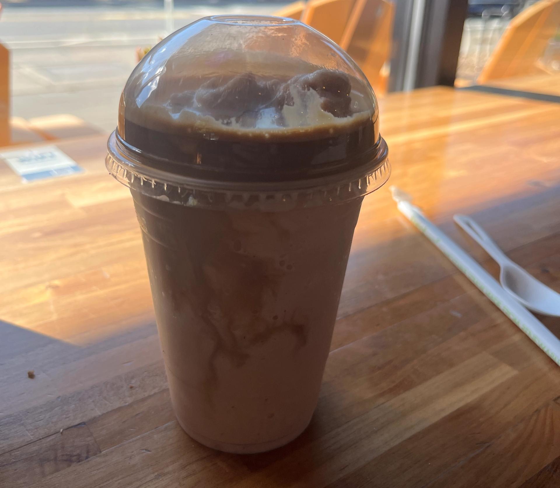 An iced coffee drink with swirls of cream in a clear plastic cup with a domed lid, sitting on a wooden table by a sunny window.