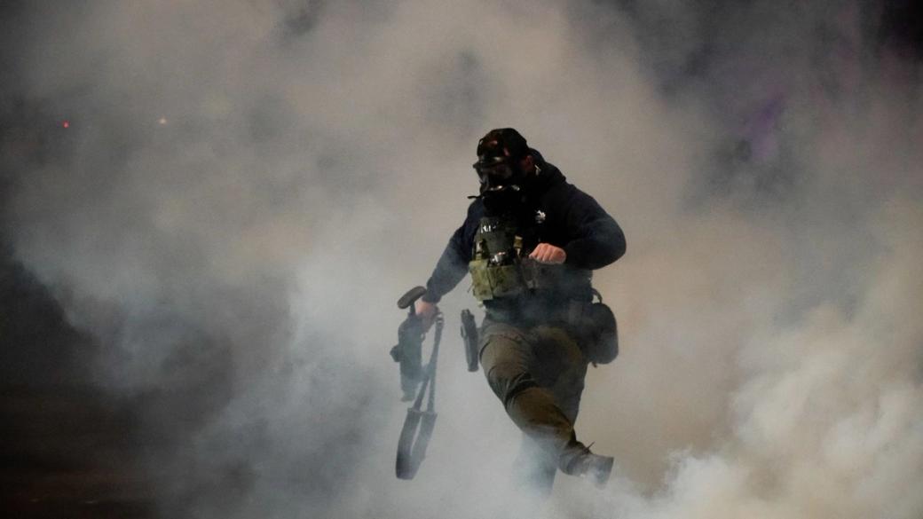 A federal agent wearing a gas mask and holding a rifle is surrounded by a cloud of white smoke. His right leg is in the air as he has just kicked a canister of tear gas toward protesters.