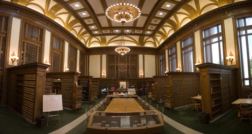 The Law Library is wood and while walls with gold gilding. There are two golden chandeliers hanging from the ceiling and bookcases with hardcover books on the left and right. A large table is in the center of the room.
