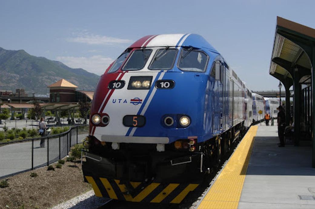 Front of the red, white, and blue FrontRunner train pulled up at a station with mountains in the background. 
