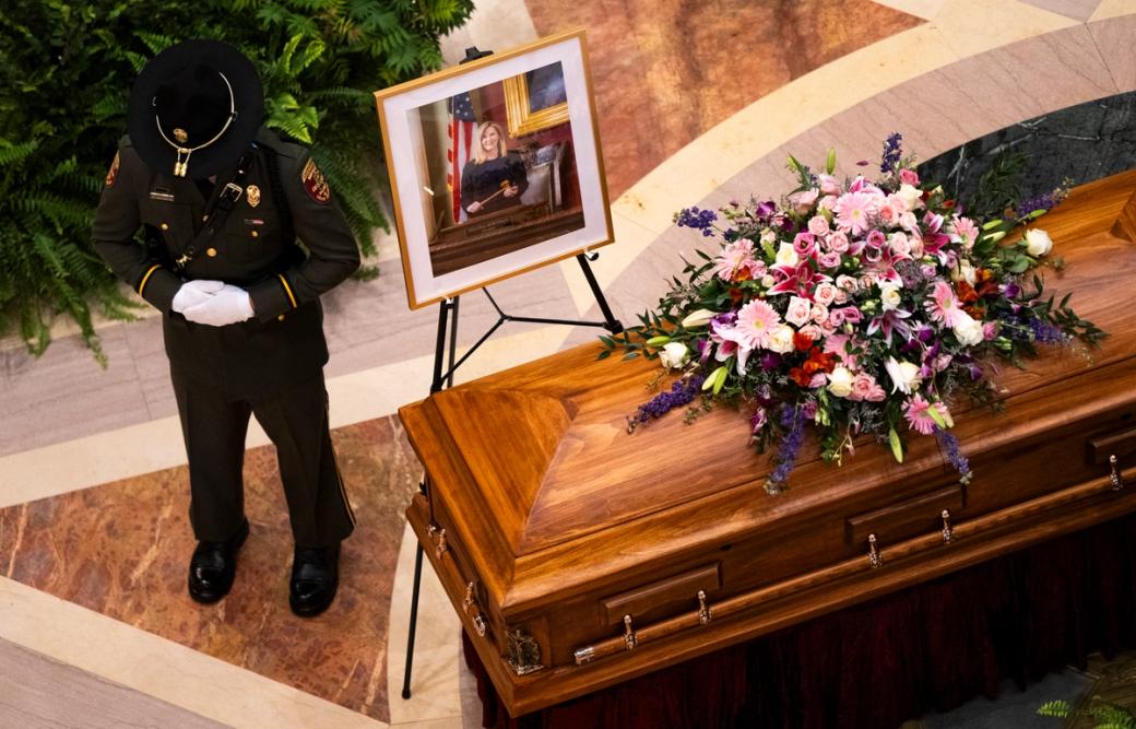 A Minnesota Conservation Officer stands guard as DFL Rep. Melissa Hortman, her husband Mark Hortman, and their dog Gilbert lie in state in the rotunda of the Capitol building. A flower-topped coffin is to the right of the guard, and behind it is a framed photo of Rep. Hortman. 