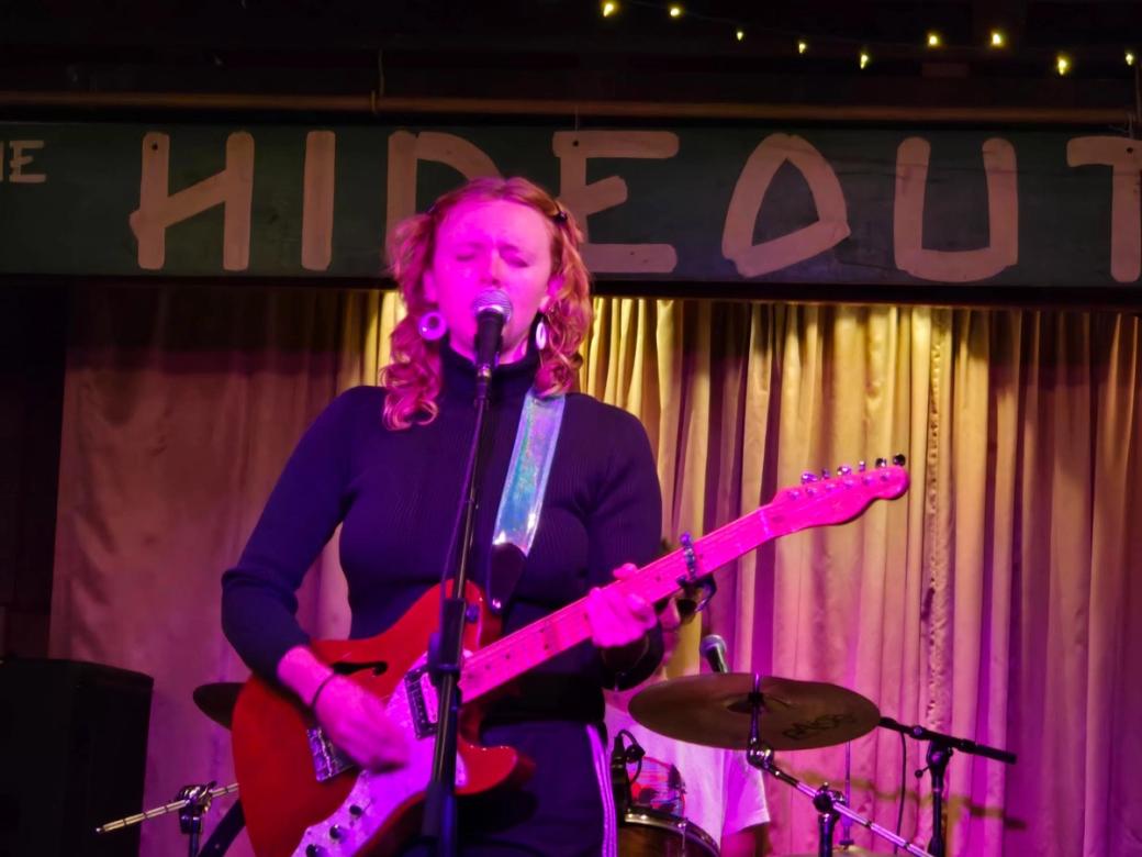 A person with a guitar on a stage in purple lighting singing into a mic