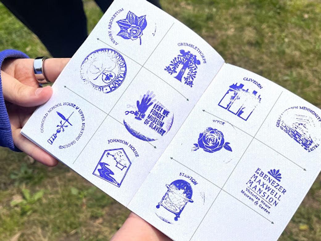 A white hand holds a booklet with purple stamps depicting the logos of several historic sites in Germantown. 