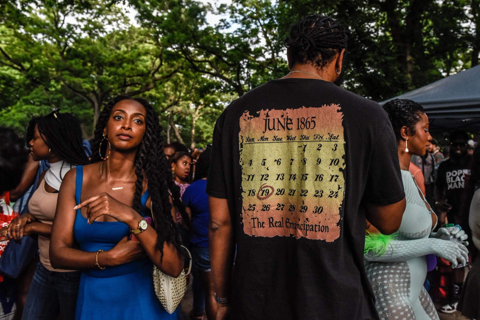 a woman with long hair faces the camera next to a man in a tshirt with his back to the camera. the shirt shows a calendar for 1865
