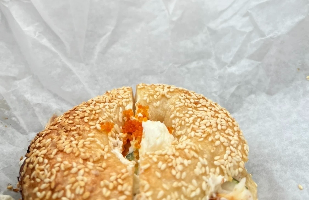 A photo of a sesame bagel with fish roe and cream cheese visible through the hole in the center, set against wax paper