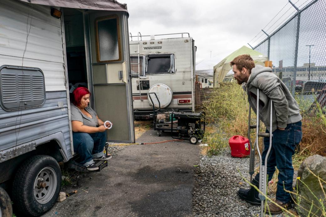 A person with dyed red hair holds a metal washer while sitting on the steps of her RV. A man on crutches stands across from her mid-conversation. Behind them another RV is parked along a chain link fence.