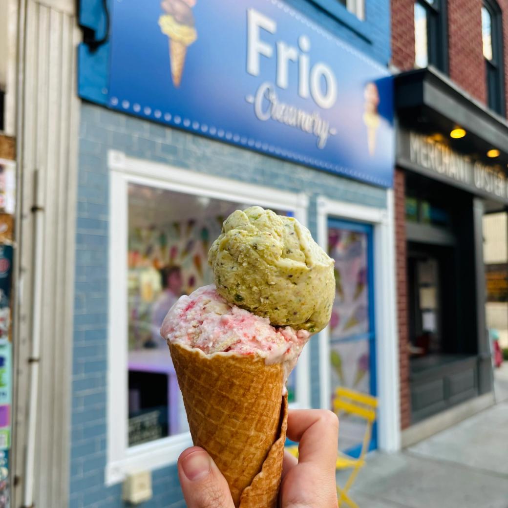 A waffle cone with a scoop of pink and creamy strawberry ice cream and a scoop of green pistachio ice cream on top in front of the blue "Frio Creamery" storefront