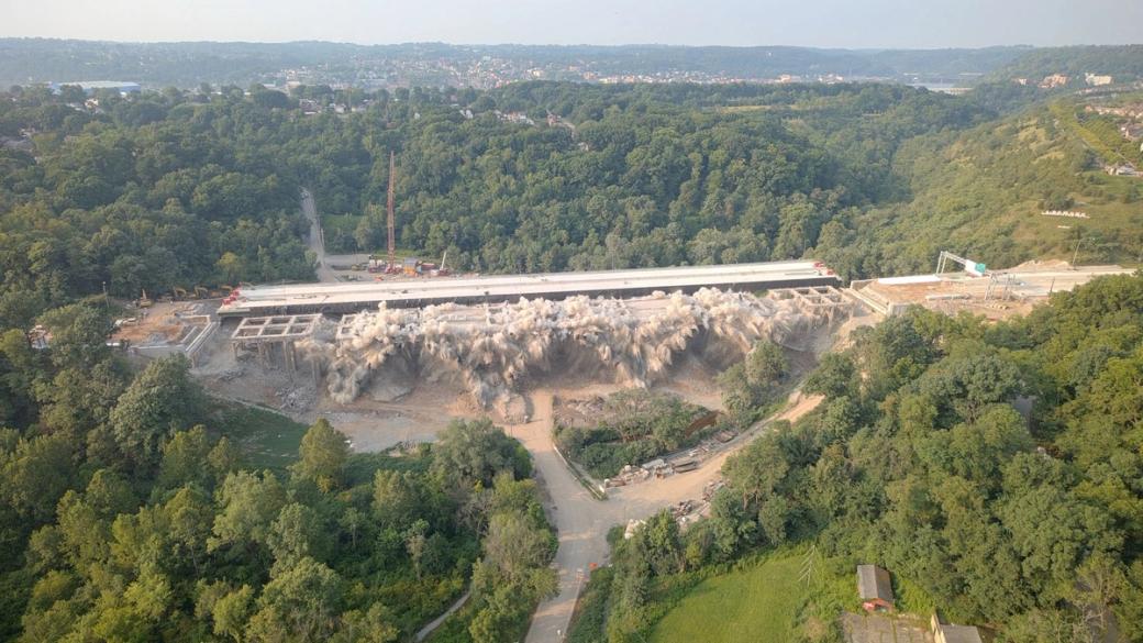 overhead view of a bridge demolition, the bridge is midway through an explosion surrounded by green grass and trees