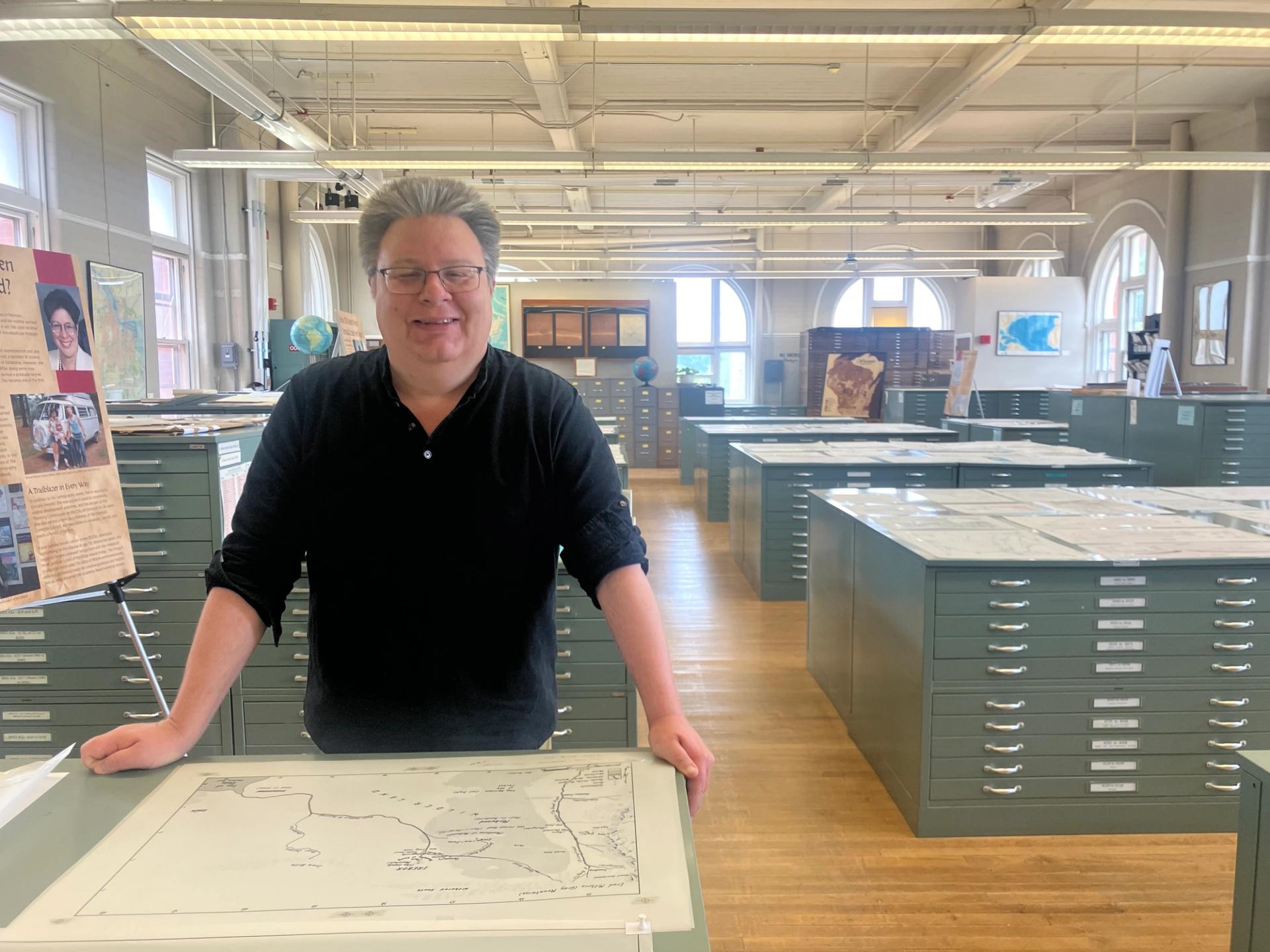 A man in a black shirt stands among tables covered in maps.