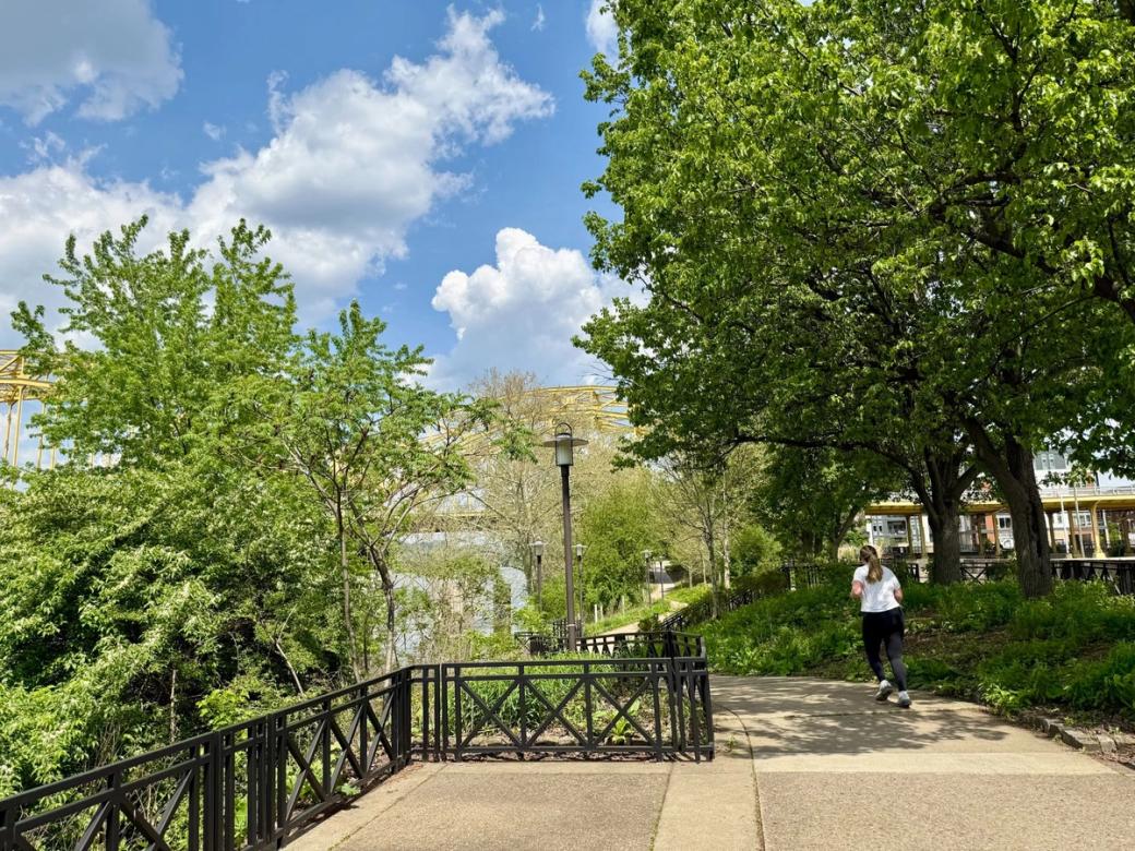 A paved riverfront trail winds through lush green trees and landscaping, with a person running along the path and a yellow bridge visible through the trees beneath a bright blue sky.