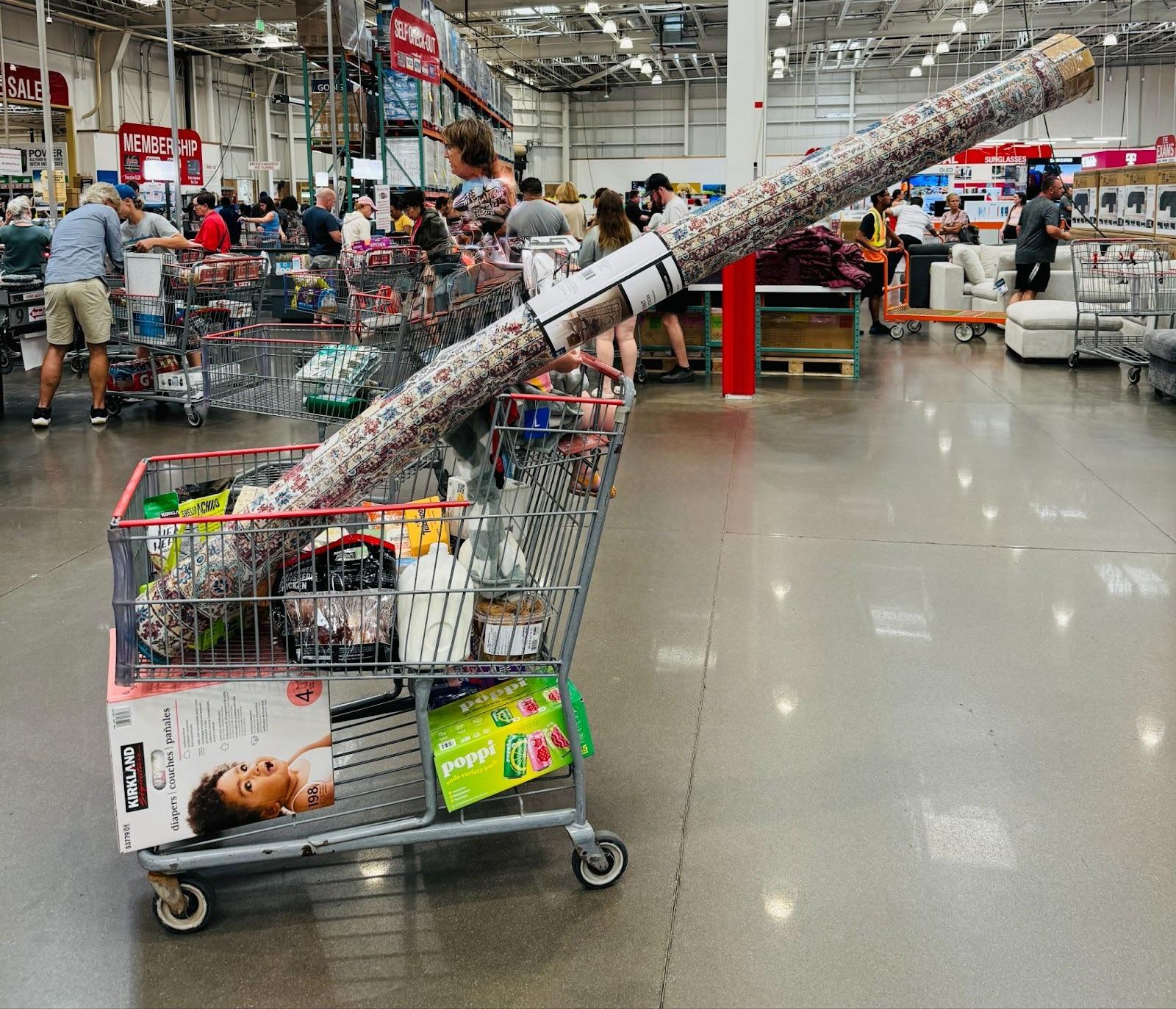 A shopping cart filled with food items and a long rolled up rug that does not fit and hangs over where the person would be pushing it