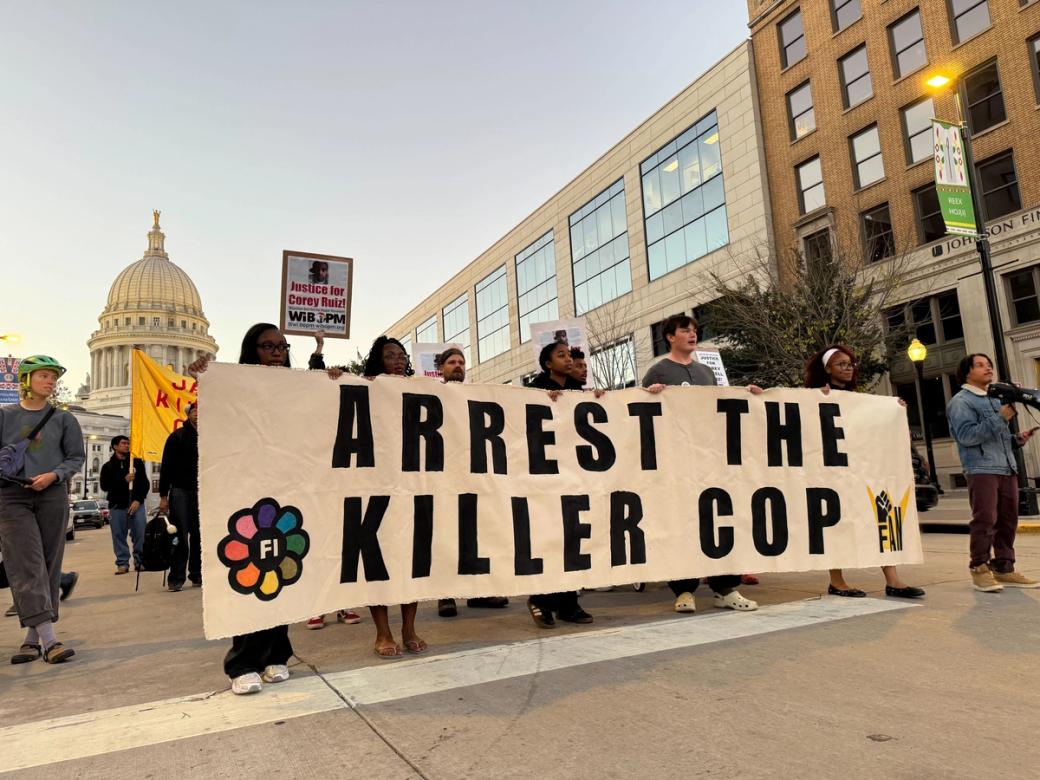 A small group of protestors stand holding a sign that says "arrest the killer cop" in front of the Wisconsin State capitol.
