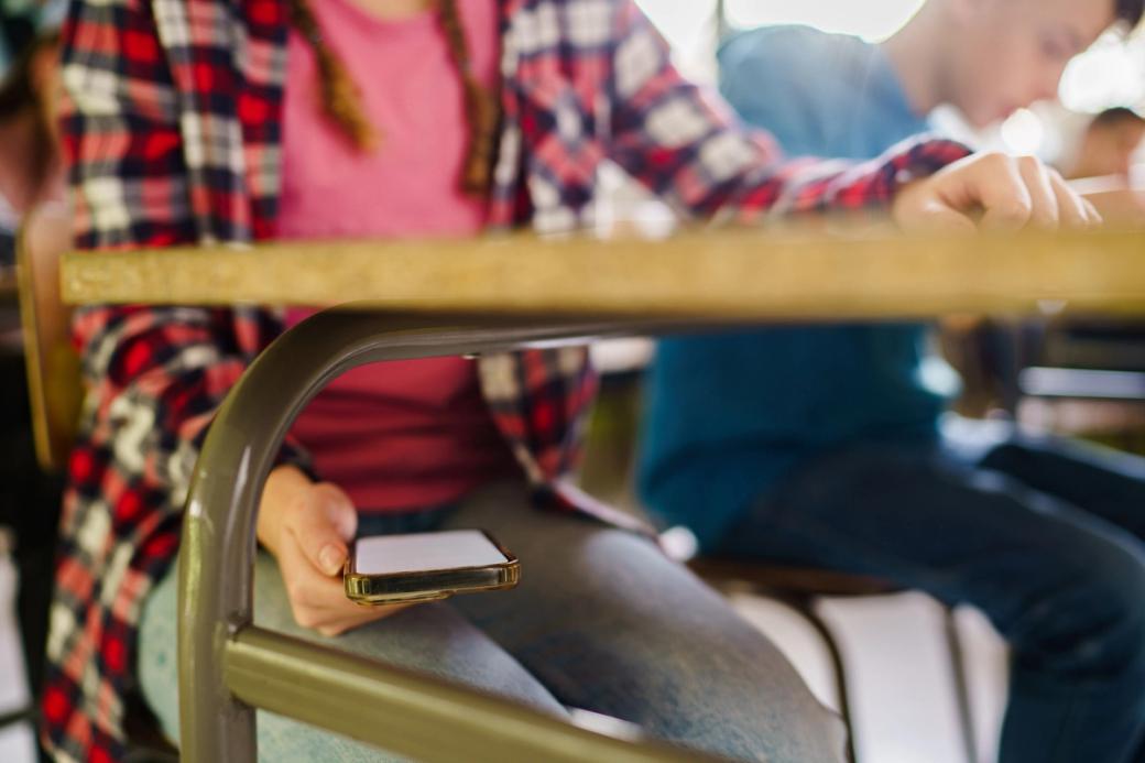 A student secretly holds a smartphone under a school desk while classmates work around them.