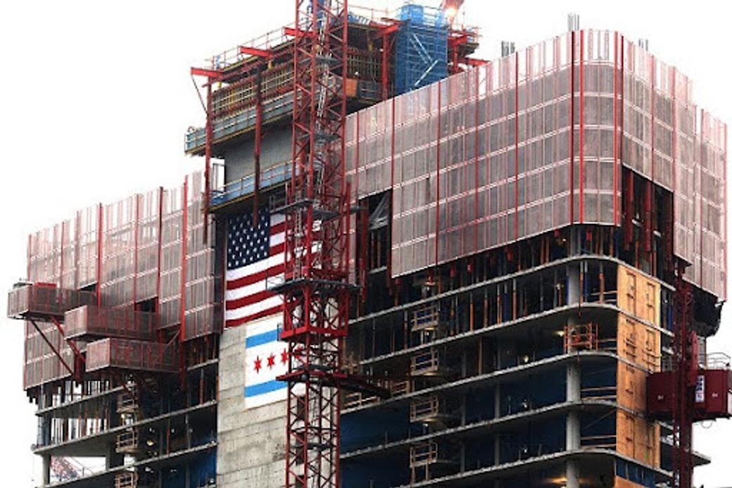 A high-rise building under construction with red scaffolding, displaying a large American flag alongside a Chicago city flag on the concrete facade.