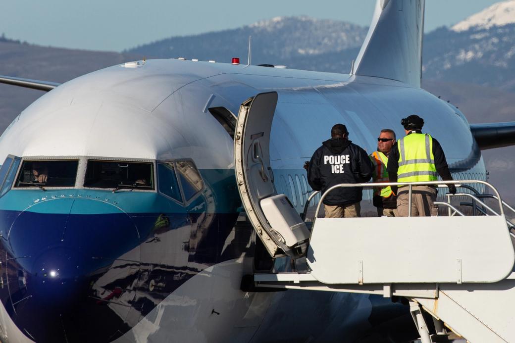 A plane sits on the tarmac with three men, two in high-vis vests and one wearing a jacket reading “Police ICE” in white block letters, standing at the opening to the plane in mid-conversation.